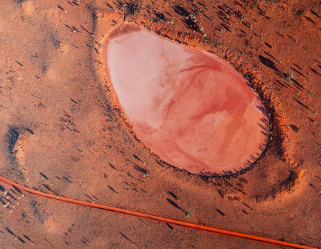 Aerial view of a vibrant red salt lake surrounded by arid outback terrain with sparse vegetation and a long dirt road in Australia.