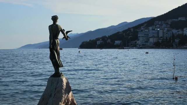 A scenic video of the iconic Maiden with the Seagull (Djevojka s galebom) sculpture standing on a rocky outcrop along the Lungomare waterfront in Opatija, Croatia. Kvarner Bay coastline appear in the 