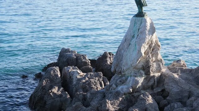 A scenic video of the iconic Maiden with the Seagull (Djevojka s galebom) sculpture standing on a rocky outcrop along the Lungomare waterfront in Opatija, Croatia