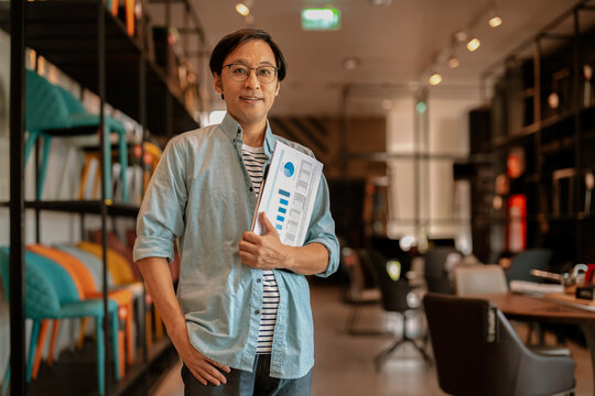 Confident Asian business owner smiling at camera holding project report chart in modern furniture showroom, demonstrating professional competence, friendly leadership, and retail store management.