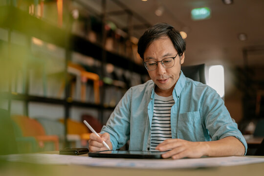 Focused Asian architect drawing on digital tablet with stylus at office desk under warm ambient light, demonstrating concentrated diligence, creative drafting precision, thoughtful planning, absort