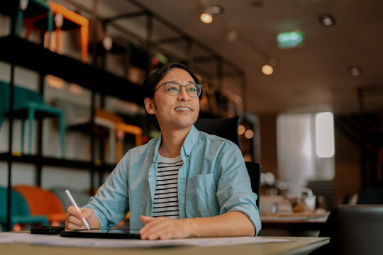Focused Asian architect drawing on digital tablet with stylus at office desk under warm ambient light, demonstrating concentrated diligence, creative drafting precision, thoughtful planning, absort