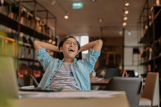 Happy Asian freelancer relaxing comfortably with hands behind head at modern coworking desk, demonstrating career satisfaction, employee wellbeing, contented productivity, work-life balance, and freed