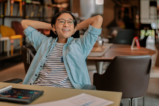 Happy Asian freelancer relaxing comfortably with hands behind head at modern coworking desk, demonstrating career satisfaction, employee wellbeing, contented productivity, work-life balance, and freed