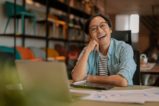 Happy Asian freelancer relaxing comfortably with hands behind head at modern coworking desk, demonstrating career satisfaction, employee wellbeing, contented productivity, work-life balance, and freed