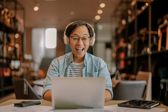 Excited Asian freelancer wearing headphones celebrating victory during video call on laptop at modern workspace, demonstrating virtual meeting success, enthusiastic achievement, and remote breakthroug