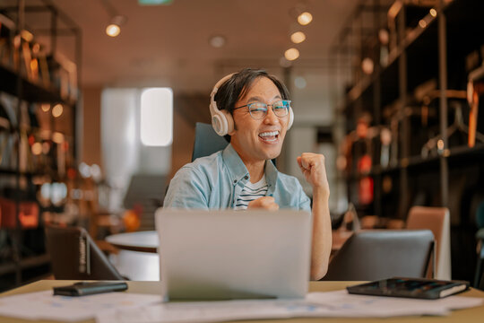 Excited Asian freelancer wearing headphones celebrating victory during video call on laptop at modern workspace, demonstrating virtual meeting success, enthusiastic achievement, and remote breakthroug