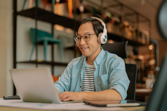 Excited Asian freelancer wearing headphones celebrating victory during video call on laptop at modern workspace, demonstrating virtual meeting success, enthusiastic achievement, and remote breakthroug