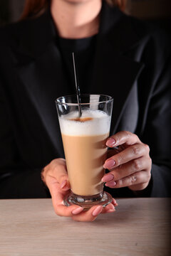 Beautiful female hands are holding a tall glass with a coffee drink. A girl drinks a latte. Latte coffee with foaming milk in a tall glass. Dark background.