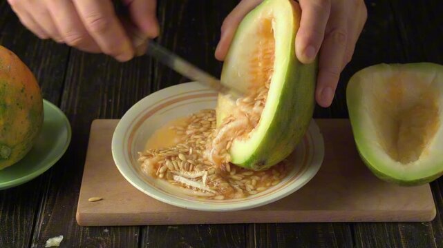 Hands using a knife to scoop out seeds from a halved melon, with a bowl of seeds and two melon halves on a wooden surface, showcasing the process of preparing fresh fruit for consumption