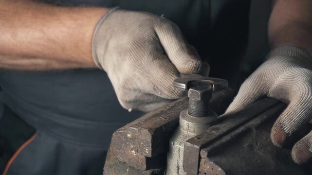 Skilled technician in gray work gloves tightens a bolt using a wrench on a metal vice, showcasing a detailed view of hands and tools in a workshop environment with industrial elements