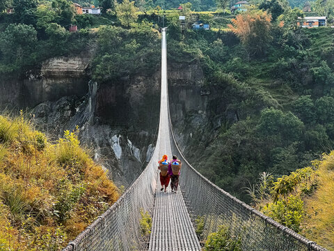 Two Nepalese women with heavy water baskets walking across a long suspension footbridge over a deep canyon in Pokhara, Nepal