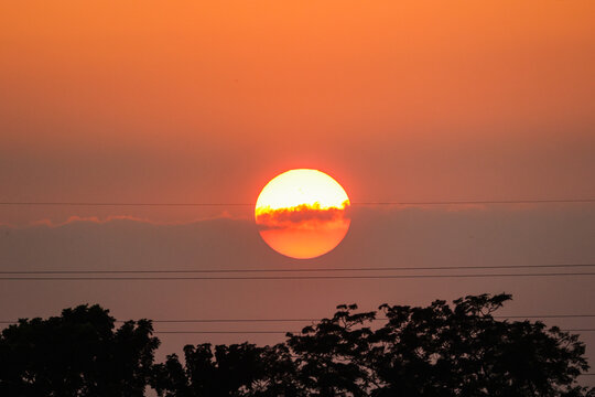 View of the sun setting behind clouds and power lines above tree silhouettes in Dhaka, Dhaka Division, Bangladesh.