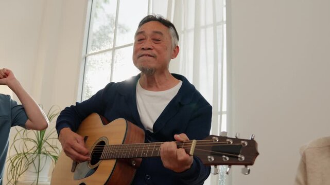 Elderly senior retirement lifestyle man singing guitar music indoors sunlit window, playing acoustic guitar in bright room with joyful expression, calm home leisure scene and warm melody
