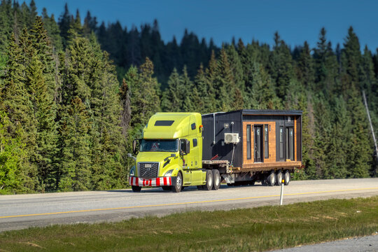 Canmore, AB, Canada - Sept 14, 2026: Yellow semi-truck transporting a pre-fabricated modular tiny house along a mountain highway.