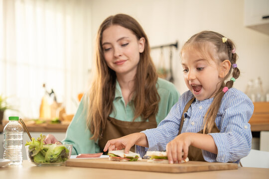Happy little girl talking excitedly to mother while sitting at kitchen table with food. Cheerful daughter sharing funny moment with smiling mother during healthy meal preparation in home kitchen.