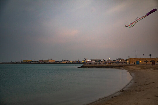View of a kite flying over the calm waters of the Persian Gulf at a beach in Doha, with distant city buildings and a sandy shoreline in Doha, Doha Municipality, Qatar.