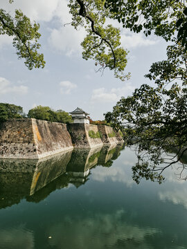 View of the historic stone walls and moat surrounding Osaka Castle with lush trees and calm water reflections in Osaka, Osaka, Japan.