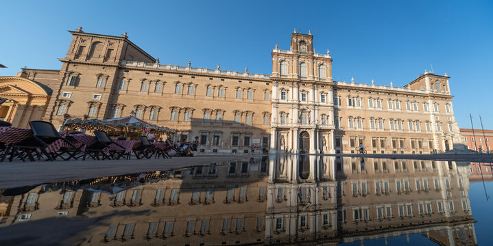 Aerial view of Palazzo Ducale di Modena with its grand facade reflected in a pool of water and a carousel on the left, Modena, Emilia-Romagna, Italy.