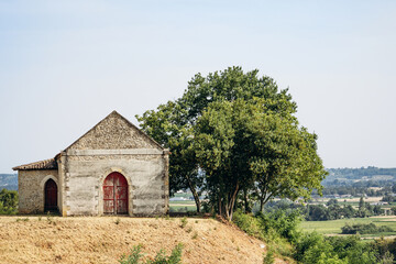 Naklejka premium Rustic stone farm building with red wooden doors and trees in the vineyard countryside near Saint Emilion, Bordeaux wine region, France.