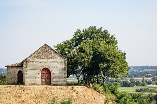 Rustic stone farm building with red wooden doors and trees in the vineyard countryside near Saint Emilion, Bordeaux wine region, France.
