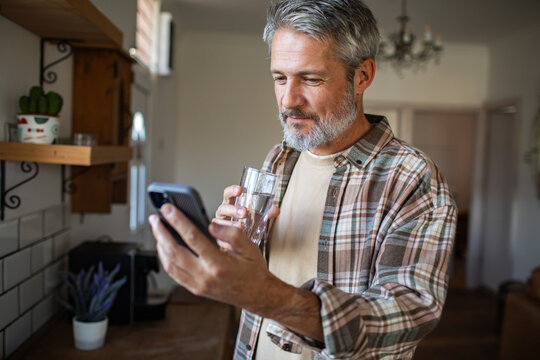 Mature man drinking water while using smartphone in home kitchen