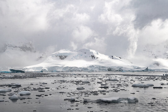 View of a snow-covered mountain landscape with floating ice chunks in the water under a cloudy sky in Antarctica.