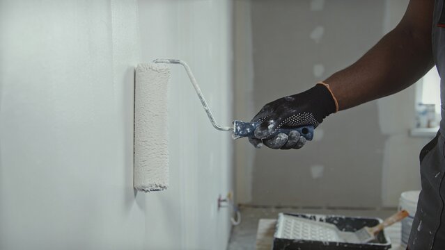 Professional painter applying fresh white interior wall coating with roller during home renovation project Skilled worker wearing protective glove carefully painting smooth surface in unfinished room 