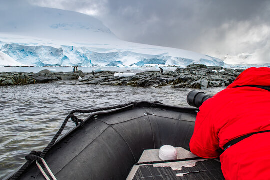 View of a photographer in a red jacket in an inflatable boat capturing images of penguins on a rocky coastline with a glacier in the background in Antarctica.