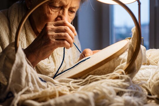 Elderly woman skillfully embroidering with blue yarn on fabric stretched in a wooden hoop under warm lamp light