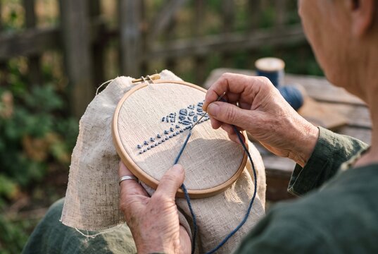 Senior Woman Hands Embroidering Blue Pattern on Linen Fabric with Wooden Hoop Outdoors