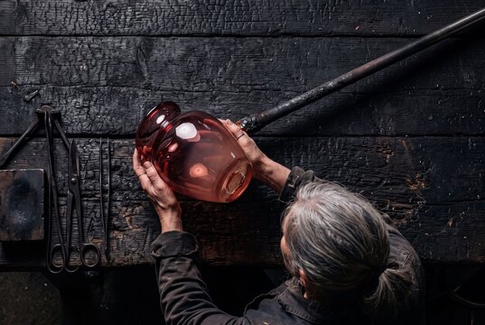 Glassblower Shaping and Inspecting Handcrafted Glass Vase at Charred Wooden Workshop Table