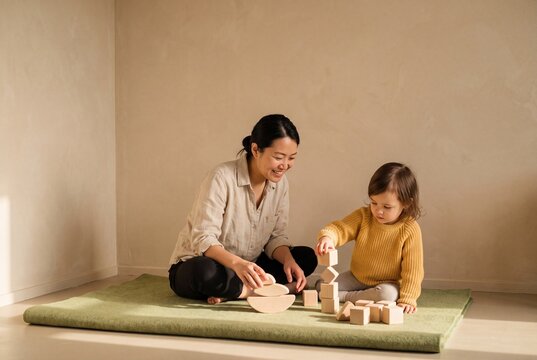 Asian mother and cute toddler daughter playing together with natural wooden building blocks on floor mat at home