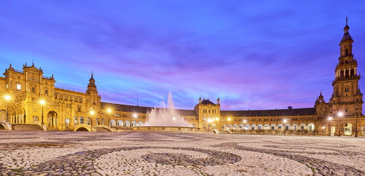 View of the illuminated Plaza de Espana in Seville with its central fountain, ornate historic architecture, and cobblestone square under a vibrant twilight sky, Andalusia, Spain.