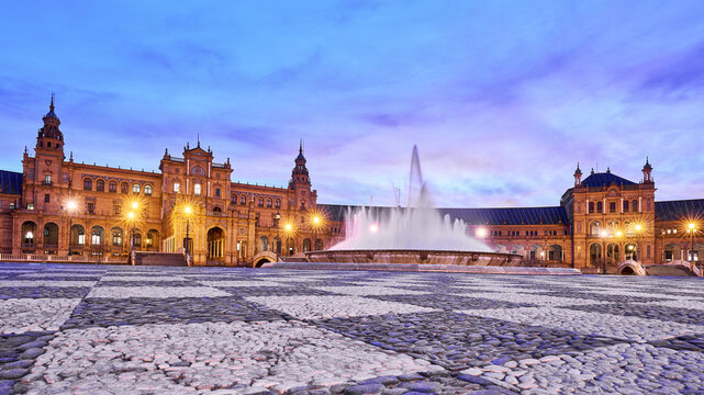 View of the illuminated Plaza de Espana with its central fountain and historic architecture under a twilight sky in Seville, Andalusia, Spain.