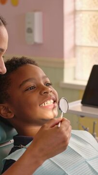 Vertical video: Leaning dentist guiding child with bib checking teeth with mirror in seat for check