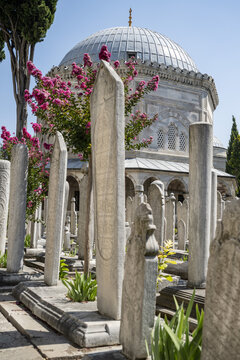 View of historic stone tombstones in a cemetery with a domed mausoleum in the background and blooming crape myrtle trees under a clear blue sky in Istanbul, Istanbul, Turkiye.