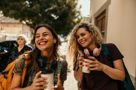 Young women friends laughing walking city street enjoying coffee