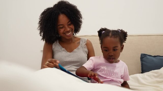 Happy african american mother and her little daughter reading a book together in bed. Loving family moment enjoying a bedtime story at home