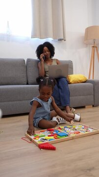 African american mother working from home on her laptop and talking on the phone while her little daughter plays with a wooden puzzle on the floor