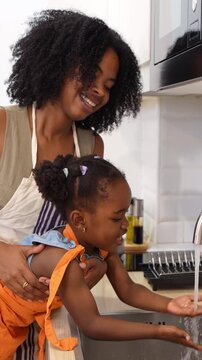 Joyful african american mother helping her little girl wash her hands in the kitchen sink. A happy family moment promoting hygiene and health
