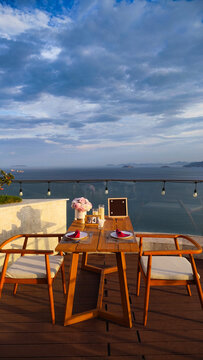 Empty wooden table and chairs setup on a rooftop cafe balcony overlooking a calm blue sea, coastal mountain landscape, and green tropical plants under a partly cloudy sky. Perfect background for