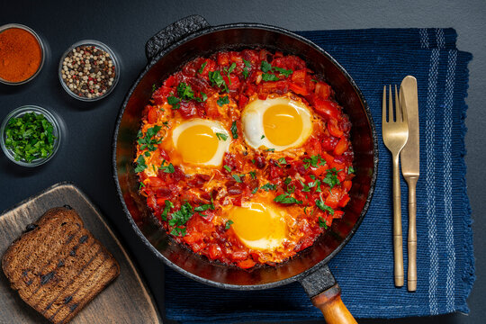 Flat lay of traditional Middle Eastern shakshuka, poached eggs in spicy tomato sauce, pepper and onion, garnished with fresh parsley, closeup. Cozy restaurant breakfast food background
