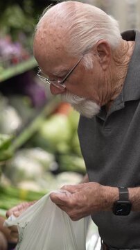 An elderly man is shopping in a grocery store. He selects vegetables and puts them in a plastic bag as he shops.