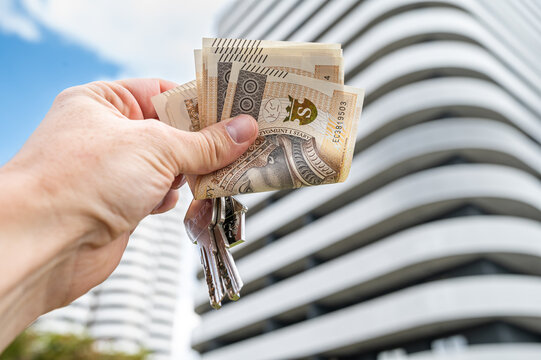 Male Hand Holding House Keys with 200 Polish Zloty Banknotes Against Modern Apartment Building