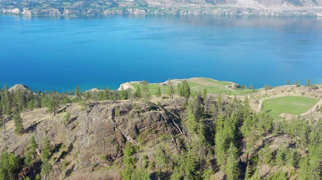 Aerial view over a rocky hill revealing vineyard plantations and Okanagan Lake shoreline in British Columbia, Canada A scenic aerial drone shot flying over rugged rocky bluffs and pine trees