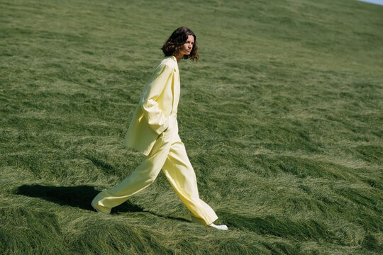 Side profile of a model in pale yellow oversized suit walking across a windswept green hill for fashion background and banner design