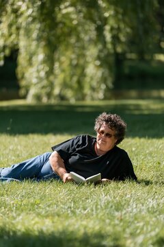Middle-aged man lying on grass reading a book with sunglasses in a sunlit park for relaxation portrait and lifestyle background use