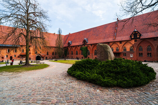 Malbork, Poland - 23 March 2026: View of the historic Malbork Castle courtyard with its red brick architecture, cobblestone ground, and a large stone monument surrounded by greenery.