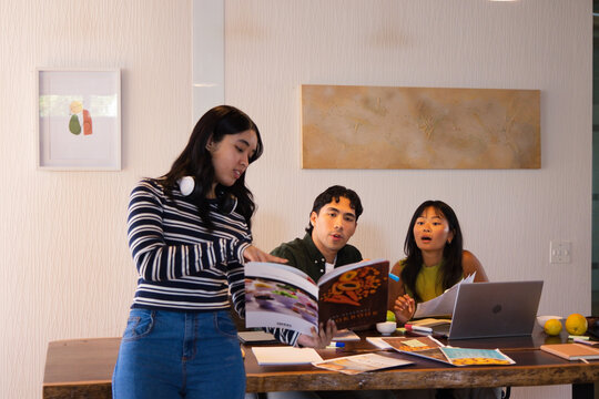 Diverse students leaning in, discussing project at dining table with laptop, open book, headphones